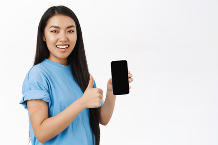 Smiling korean woman with bandaid after coronavirus vaccine, shows smartphone screen, recommends covid health certificate on mobile phone, white backgroundの写真素材