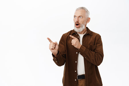Surprised old guy, mature bearded man with grey hair, pointing fingers left, looking at copy space with impressed face expression, standing over white backgroundの写真素材