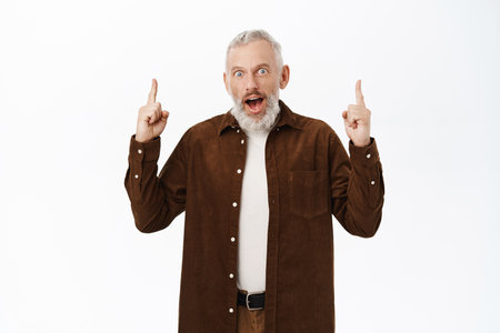 Enthusiastic senior man, mature guy wtih grey hair, pointing fingers up, showing advertisement on top, standing over white backgroundの写真素材