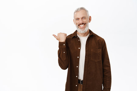 Smiling senior man with grey beard and white smile, pointing finger left, showing website, logo or banner, standing against studio backgroundの写真素材