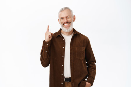 Image of smiling handsome bearded man with grey hair, senior guy pointing finger up, showing advertisement, standing over white backgroundの写真素材