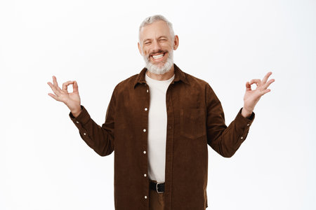 Mindfulness and relaxation concept. Senior man, old guy meditating, holding hands in namaste zen gesture, standing over white backgroundの写真素材