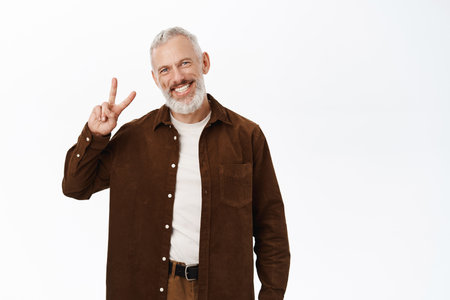 Handsome senior man, old male model smiling showing peace, v-sign gesture, standing carefree in casual clothes against studio backgroundの写真素材