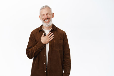Image of senior guy, old man being touched, holding hands on chest grateful, looking flattered and pleased, standing over white backgroundの写真素材