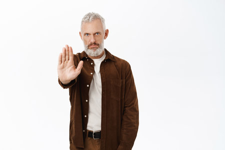 Portrait of serious senior man, mature male model extending palm, showing stop, prohibit gesture, standing over white backgroundの写真素材