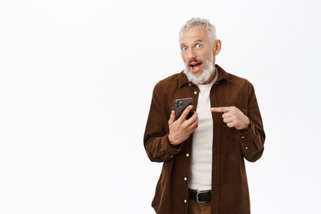 Handsome old man, dad with grey hair, pointing finger at mobile phone, using smartphone app with happy face, white backgroundの写真素材