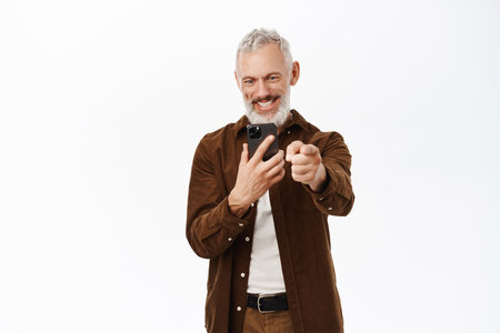Senior man, mature guy pointing finger at camera and recording video, taking pictures on smartphone, standing over white backgroundの写真素材