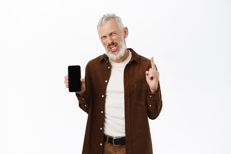 Portrait of senior man, old caucasian guy showing mobile phone screen, pointing finger up, standing in casual clothes over white backgroundの写真素材