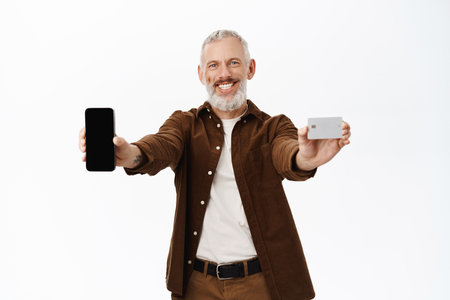 Handsome senior man showing mobile phone screen, app interface and credit card, standing over white backgroundの写真素材