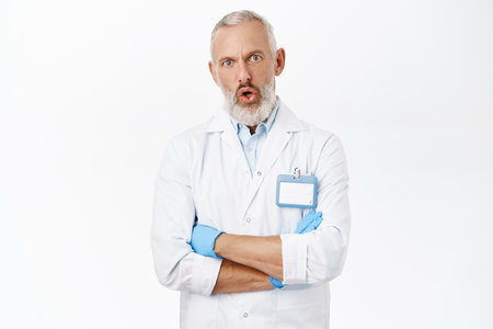 Shocked senior doctor in white coat, looking confused and startled at camera, posing against white backgroundの写真素材