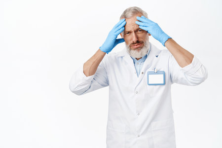 Tired senior male doctor, holding hands on head, migraine, distressed after work hospital, standing against white backgroundの写真素材