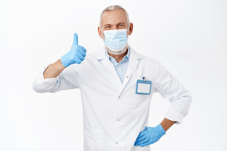 Doctor in medical face mask from covid-19, wearing rubber gloves and white coat, showing thumbs up, recommending hospital or clinic, standing over white backgroundの写真素材