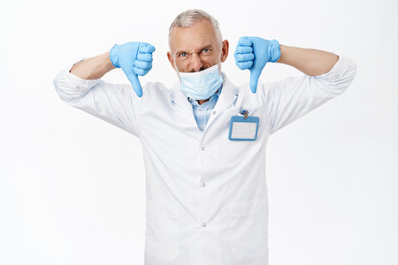 Coronavirus and health concept. Angry doctor in medical face mask, showing thumbs down dislike gesture, disapprove smth, standing over white backgroundの写真素材