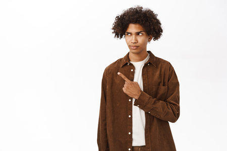 Portrait of skeptical young man, pointing and looking left, staring with doubtful face expression, standing over white backgroundの写真素材