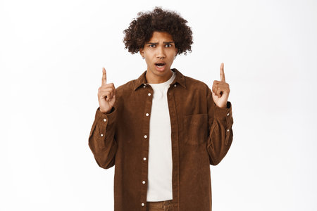 Shocked guy pointing fingers up, looks worried and concerned, stands over white backgroundの写真素材