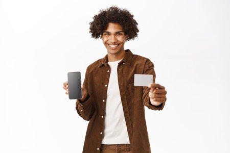Portrait of smiling hispanic man shows mobile phone screen and credit unior card, advertisement of bank, standing over white backgroundの写真素材