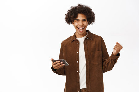 Image of happy young hispanic boy holding mobile phone and triumphing, celebrating good news, standing over white backgroundの写真素材