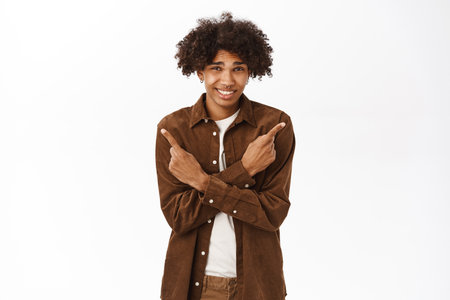 Portrait of smiling gen z guy with afro hair, pointing fingers sideways, showing left and right side, logo or product banners, give choice, standing over white backgroundの写真素材