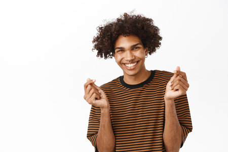 Close up of handsome curly guy shows finger hearts and smiling cute at camera, standing over white backgroundの写真素材