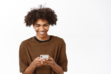 Portrait of young curly guy chatting on smartphone app, using mobile phone and looking at screen with happy smile, standing over white backgroundの写真素材