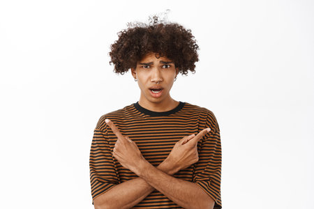 Portrait of young guy with confused face pointing sideways, cant decide which side to choose, standing over white backgroundの写真素材