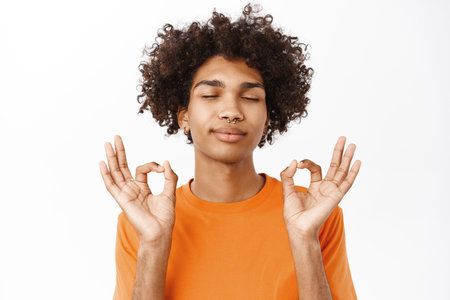 Mindfulness and meditation. Young man meditating, feeling zen and relaxtion, close eyes and showing mudra gesture, standing over white backgroundの写真素材