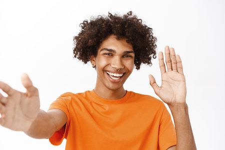 Close up portrait of hispanic guy video chat, holding mobile phone and waving at camera, laughing and smiling, standing over white backgroundの写真素材