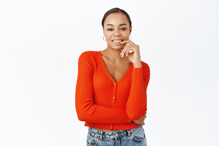 Portrait of beautiful black woman in red blouse, smiling and looking confident, interested in something, standing over white backgroundの写真素材