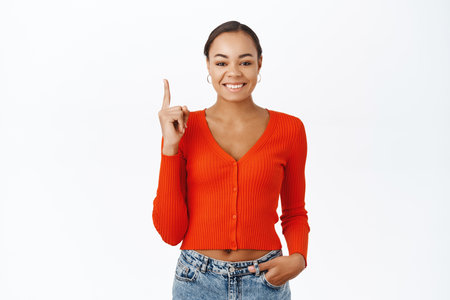 Portrait of smiling beautiful black woman, pointing finger up, showing advertisement, store discount on top, standing over white backgroundの写真素材
