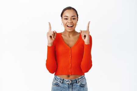 Enthusiastic young latina woman, pointing finger up, smiling and laughing with excitement, standing over white backgroundの写真素材