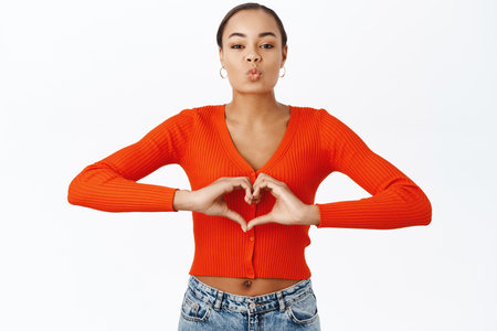 Portrait of smiling latina girl showing heart, care sign, standing in red blouse over white backgroundの写真素材