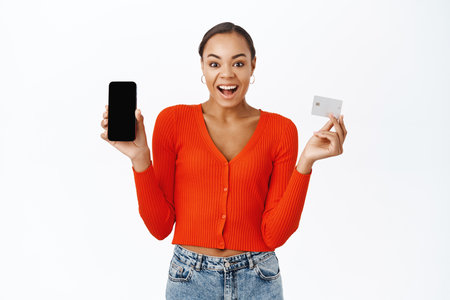 Happy young woman shows her mobile phone screen and credit card, smiling with excitement, stands over white backgroundの写真素材