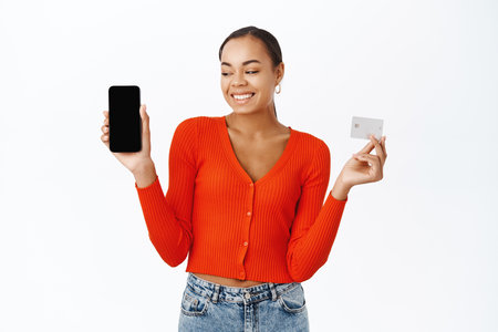 Happy young woman shows her mobile phone screen and credit card, smiling with excitement, stands over white backgroundの写真素材