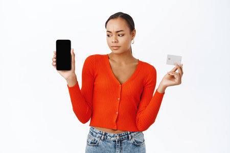 Disappointed woman showing credit card, looking upset at mobile phone screen app, frowning upset, standing over white backgroundの写真素材