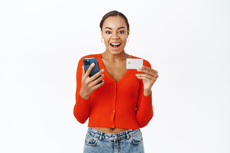 Technology and online shopping. Smiling woman using mobile phone and credit card, standing happy over white background, ordering smthの写真素材