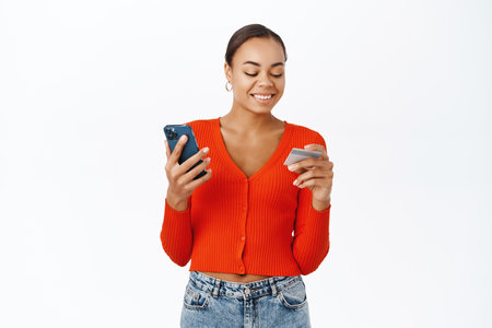 Portrait of beautiful young woman using credit card and smartphone, making order at online store, purchase smth in application, standing over white backgroundの写真素材
