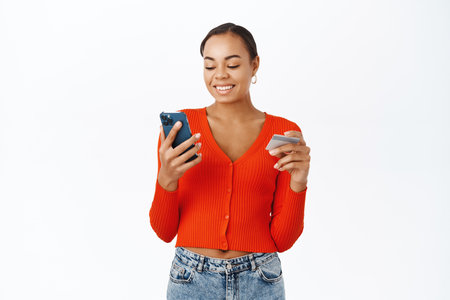 Portrait of beautiful young woman using credit card and smartphone, making order at online store, purchase smth in application, standing over white backgroundの写真素材