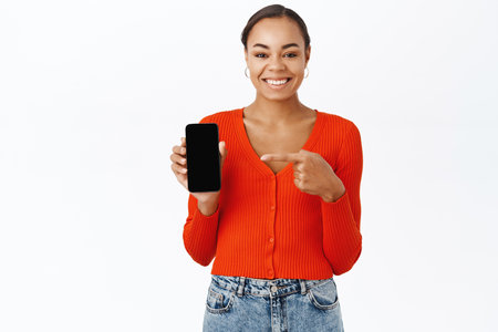 Portrait of woman pointing finger at mobile phone screen, showing smartphone application, standing over white backgroundの写真素材