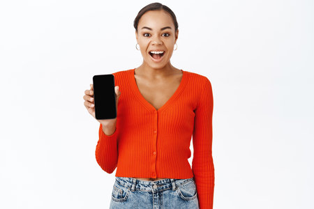 Enthusiastic young woman smiles, shows smartphone screen, mobile application, stands over white studio backgroundの写真素材