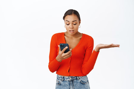 Woman looks confused at smartphone screen, shrugs shoulders and complains at smth on mobile phone, stands over white backgroundの写真素材