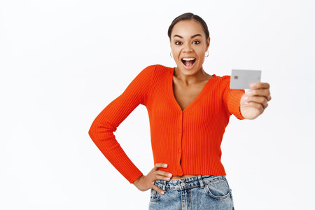 Smiling young woman showing credit card, recommending bank services, posing in red blouse against white backgroundの写真素材