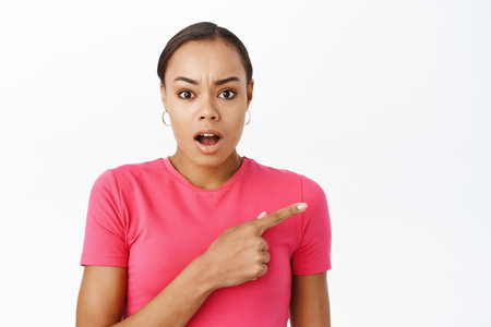 Close up of surprised woman points finger right, gasps and looks shocked, impressed by smth, stands over white backgroundの写真素材