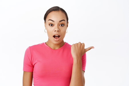Close up of impressed female model, points finger right, gasps with wow face, amazed by smth, stands over white backgroundの写真素材