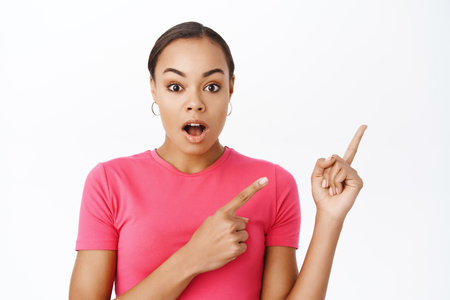 Close up of impressed female model, points finger right, gasps with wow face, amazed by smth, stands over white backgroundの写真素材