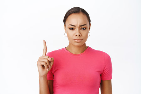 Close up of angry black woman, frowns and points up with disappointed face expression, stands over white backgroundの写真素材