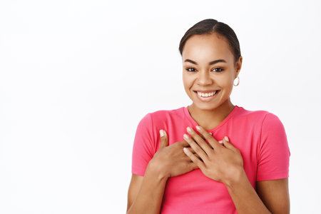 Close up of smiling woman looks flattered, holds hand on chest, feels appreciation, stands over white studio backgroundの写真素材