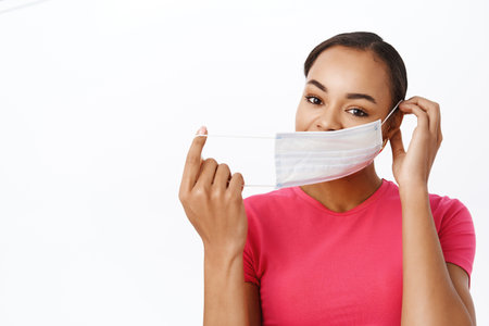 People during pandemic. Smiling black woman puts on medical face mask to protect from covid-19, standing in tshirt over white backgroundの写真素材