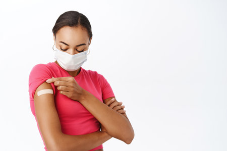 Portrait of black woman showing her shoulder with patch after covid-19 vaccination, wearing medical face mask, standing over white backgroundの写真素材