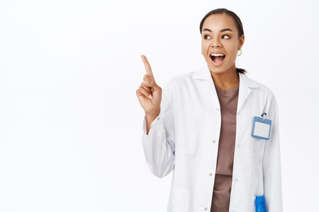 Woman doctor in white coat, pointing finger and looking at banner, showing medical clinic logo on copy space, standing over white backgroundの写真素材