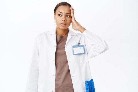 Troubled female doctor, holds hand on head, look concerned and complicated, stands against white backgroundの写真素材
